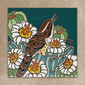 May include: A square decorative tile featuring a brown and white bird with a long tail perched among green cacti and white flowers with bright orange centers. The background is a deep teal color, and the design uses bold, dark outlines.