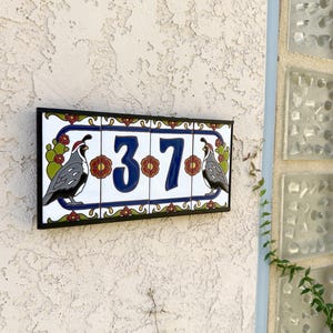 May include: A rectangular house number plaque featuring four ceramic tiles framed in black. The design displays the digits 3 and 7 in dark blue, flanked by illustrations of grey and white quail birds, green cacti, and small red floral accents.