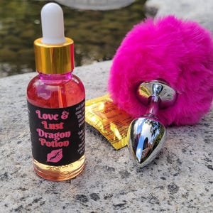 Könnte beinhalten: Ein rosa flauschiger Schwanz mit einem silbernen Metallstopfen und eine kleine Flasche mit klarer Flüssigkeit mit einem schwarzen Etikett, auf dem "Love & Lust Dragon Potion" steht.