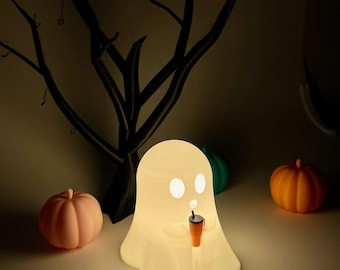 May include: A lit ghost-shaped lamp with eye holes and a small orange cup with a straw. Two small pumpkins, one pink and one orange, are on either side of the lamp. A dark tree silhouette is in the background.
