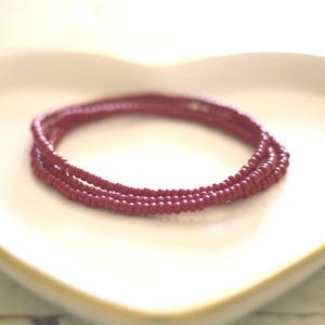 May include: A multi-strand bracelet composed of small, uniform, matte-finish dark red beads. The circular accessory features a consistent, monochromatic color scheme throughout its design, with each individual bead showing a smooth, rounded surface texture.