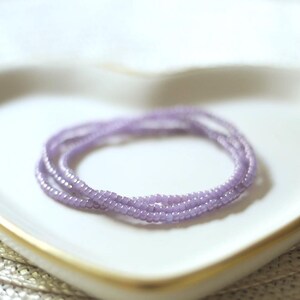 May include: A delicate bracelet composed of multiple strands of small, uniform, light purple seed beads. The beads have a subtle, smooth texture and are arranged in a circular, layered formation, creating a simple and consistent aesthetic.