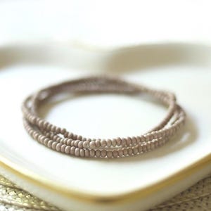 May include: A multi-strand bracelet composed of small, round, matte-finish beads in a muted taupe or light brown color. The beads are strung together in a continuous circular pattern, creating a layered appearance with a consistent, uniform texture.