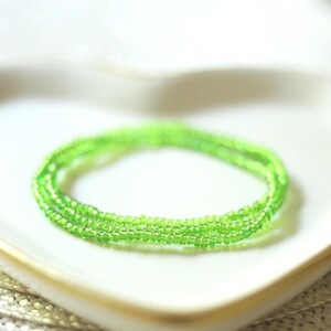 May include: A circular strand of small, translucent lime green seed beads arranged in a simple, continuous loop. The beads are uniform in size and shape, creating a vibrant, bright green ring that rests in a coiled, circular formation.