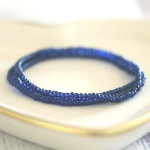 May include: A circular bracelet composed of multiple strands of small, deep blue beads. The beads have a reflective, glossy finish and are arranged in a continuous, uniform loop, creating a simple and consistent texture throughout the entire piece.
