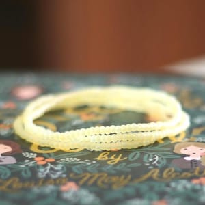 May include: A pale yellow beaded bracelet consisting of three delicate strands of small, uniform, translucent beads. The bracelet is resting on a dark green book cover featuring illustrated figures and the text Louisa May Alcott.
