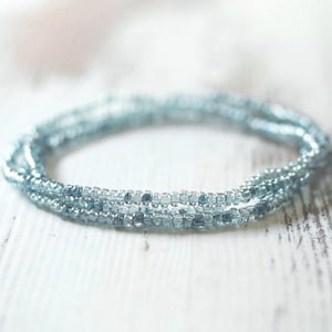 May include: A multi-strand bracelet composed of small, translucent light blue glass seed beads. The circular accessory features a consistent, uniform texture throughout its design, showcasing a delicate and reflective appearance in shades of blue.