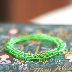 May include: A circular bracelet composed of small, vibrant green seed beads. The bracelet features a uniform, translucent appearance with a reflective surface, creating a continuous loop of bright green color that stands out clearly.