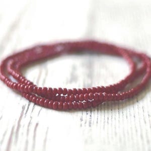 May include: A multi-strand necklace composed of small, uniform, deep red beads. The beads are round and have a smooth, matte finish, arranged in several circular loops that overlap to create a layered appearance of consistent red color.