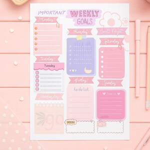 May include: A pink and purple weekly planner with sections for each day, including "Monday" through "Sunday", and a "to do list" section. The planner has decorative elements like hearts and a cloud, with pens and paperclips nearby.