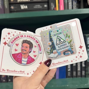 Könnte beinhalten: Zwei dekorative Karten. Eine Karte zeigt ein Porträt mit dem Text "DADDY IS A STATE OF MIND". Die andere Karte hat ein Warnschild mit dem Text "SLIPPERY WHEN READING". Beide Karten haben einen holografischen Effekt.