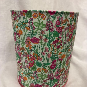 May include: A cylindrical wastebasket featuring a white background with a dense, colorful floral pattern. The design includes various small flowers in shades of pink, orange, and green, with a solid pink trim along the bottom edge.