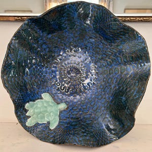 May include: A round, wavy-edged ceramic bowl featuring a deep blue and dark teal glaze with a textured, scale-like pattern across the surface. A light mint green sea turtle figure is attached to the lower left side of the interior basin.