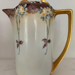 May include: A porcelain coffee pot featuring a cream-colored body with hand-painted yellow roses and green foliage. The pot includes a gold-toned handle and a matching gold-toned lid handle, with a dark brown decorative border near the top rim.
