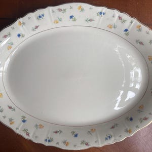 May include: Oval serving platter with a scalloped edge and a floral pattern. The white porcelain platter features a delicate border of small blue, yellow, and pink flowers, with a gold trim. A classic piece for serving.