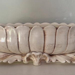 May include: Cream-colored, oval-shaped planter with a scalloped rim and ornate handles. The planter features a leaf-like base and vertical, petal-shaped sides. The aged finish adds a vintage aesthetic to this decorative piece.