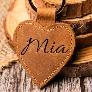 Może przedstawiać: Brązowy skórzany brelok w kształcie serca z wygrawerowanym imieniem "Mia". Brelok jest przymocowany do srebrnego kółka na klucze z wieloma kluczami.