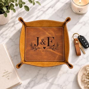 May include: A square tan leather valet tray with snap-button corners. The center is engraved with the letters J & E, a small heart, leafy branches, and the date 12.14.2024. The tray features visible white stitching along the edges and sides.