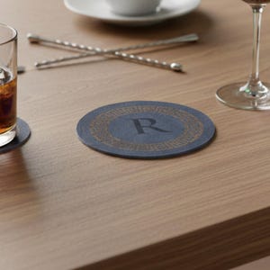 Könnte beinhalten: Ein runder, marineblauer Untersetzer mit goldfarbenem griechischem Schlüsselmuster und dem Buchstaben "R" in der Mitte. Der Untersetzer liegt auf einem hellbraunen Holztisch, im Hintergrund ein Glas mit bernsteinfarbener Flüssigkeit und ein Cocktailrührer.