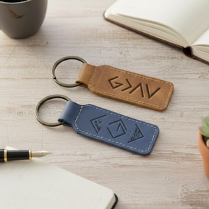 May include: Two leather keychains, one brown and one blue, with metal key rings. The brown keychain has the letters "G>AV" engraved. The blue keychain has a mountain and geometric design.
