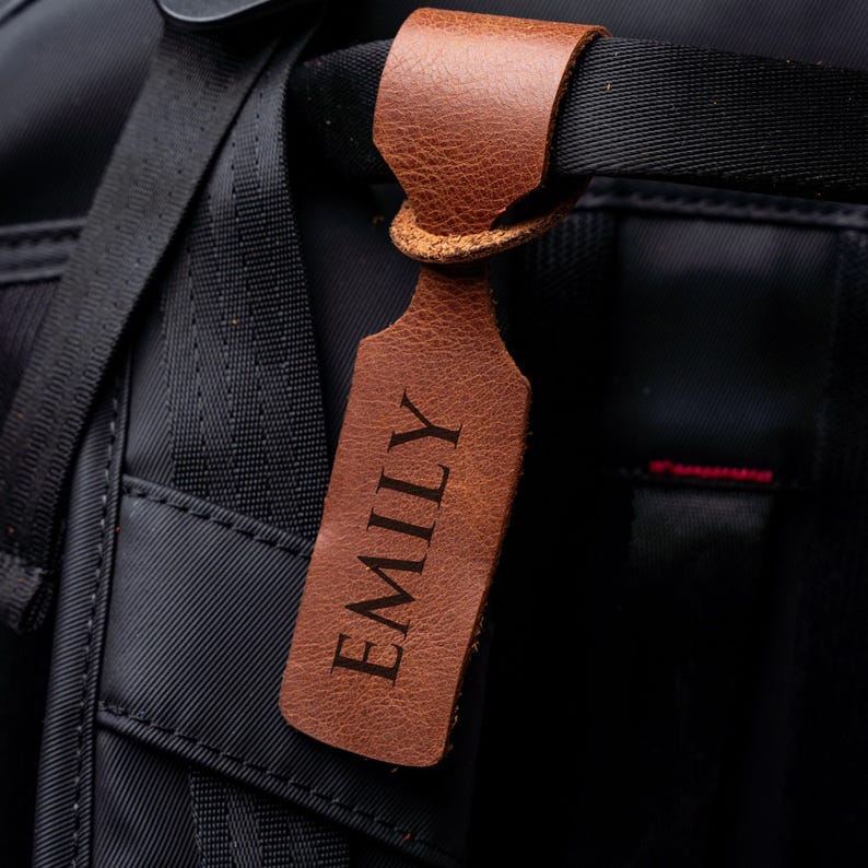 A brown leather name tag with the name "EMILY" printed on it, attached to a black strap or handle of a bag or backpack.