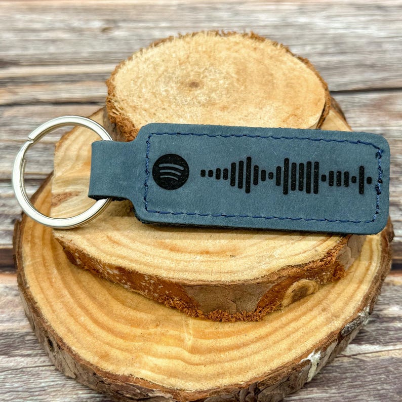 May include: A dark blue leather keychain with a black Spotify logo and a black and white barcode. The keychain is on a wooden surface.
