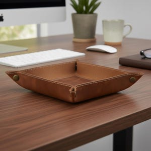 May include: A square, brown leather catchall tray with white stitching and brass-colored snap closures. The tray is empty and sits on a wooden desk, with a computer, keyboard, and mug in the background. The tray is approximately 8 inches square.