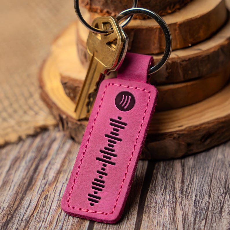 May include: A pink leather keychain with a black sound wave design and a small silver circle with three wavy lines. The keychain is attached to a silver key ring.