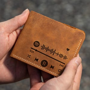 Puede incluir: Una cartera de cuero marrón con un diseño de reproductor de música grabado en la parte delantera. El diseño incluye una nota musical, un botón de reproducción, un botón de pausa y un botón de avance rápido. El texto "0:25" y "3:40" también está grabado en la cartera.