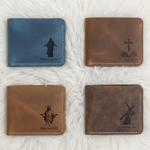 May include: Four leather wallets in shades of blue and brown are displayed on a white, fluffy surface. Each wallet features a religious image and a Bible verse. The wallets are stitched with white thread.