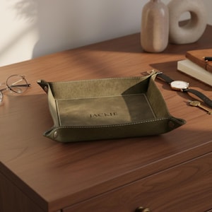 May include: Olive green leather tray with the name "JACKIE" embossed in the center. The tray has a rectangular shape with folded corners secured by metal snaps. White stitching outlines the edges. A watch, keys, and glasses are nearby.