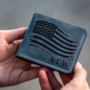 May include: A navy blue leather wallet with a black American flag design. The wallet has white stitching around the edges and the initials "ALW" are visible. The wallet is approximately 4 inches wide and 3 inches tall.