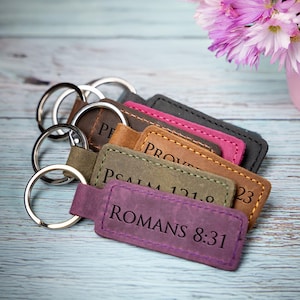 May include: A set of six leather keychains with different bible verses engraved on them. The keychains are in various colors, including brown, green, pink, and purple. The keychains are arranged in a stack, with the top keychain featuring the verse "Romans 8:31".