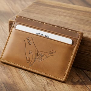 May include: A brown leather cardholder with a stitched border. The front features a drawing of two hands holding each other, with the words "DAD" and "Annie" inscribed. A credit card is visible inside the cardholder.