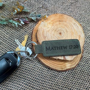 May include: Green leather keychain with the text "MATTHEW 17:20" in black lettering. The keychain is attached to a silver key ring.