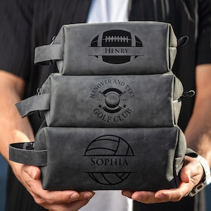 May include: Three grey leather wash bags stacked. The top bag features a football design with the name "Henry". The middle bag has a golf club design with the text "Hanover and Tyke Golf Club". The bottom bag has a volleyball design with the name "Sophia".