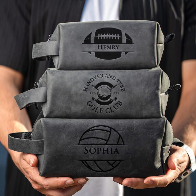 May include: Three grey leather wash bags stacked. The top bag features a football design with the name "Henry". The middle bag has a golf club design with the text "Hanover and Tyke Golf Club". The bottom bag has a volleyball design with the name "Sophia".