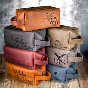 Può includere: Una pila di trousse da toilette in pelle di vari colori, tra cui marrone, grigio e bordeaux. Ogni borsa presenta una chiusura con cerniera, un manico e testo personalizzato con simboli medici e nomi come "Dr. Lucas" e "Dr. David".