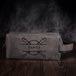 May include: A grey leather wash bag with a baseball design. The bag has a zip closure, a handle, and the name "JAMES" in a decorative font. The design includes crossed baseball bats and a baseball.