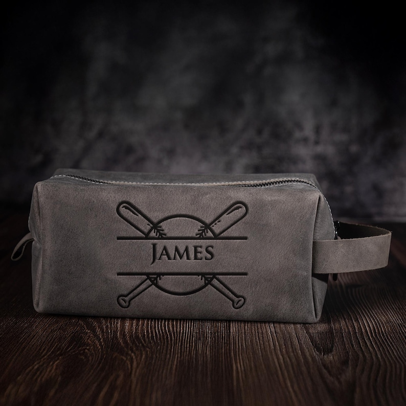 May include: A grey leather wash bag with a baseball design. The bag has a zip closure, a handle, and the name "JAMES" in a decorative font. The design includes crossed baseball bats and a baseball.