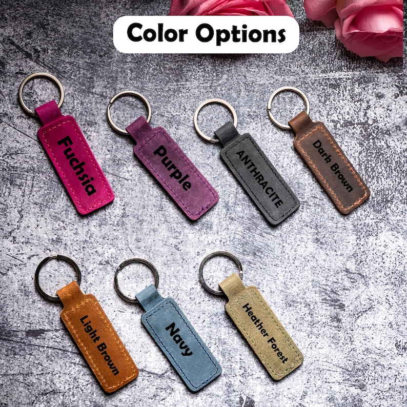May include: Seven leather keychains with different colors and engraved text. The colors are fuchsia, purple, anthracite, dark brown, light brown, navy, and heather forest.