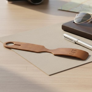 A brown leather tag with the initials 'LTM' is placed on a wooden surface, accompanied by a pen, a pair of sunglasses, and a notebook.