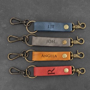 May include: Four leather keychains in blue, gray, orange, and red. Each keychain has a metal clasp and a metal ring. The blue keychain has the initials "LJT", the gray keychain has "JDH", the orange keychain has "ANGELA", and the red keychain has the letter "R".