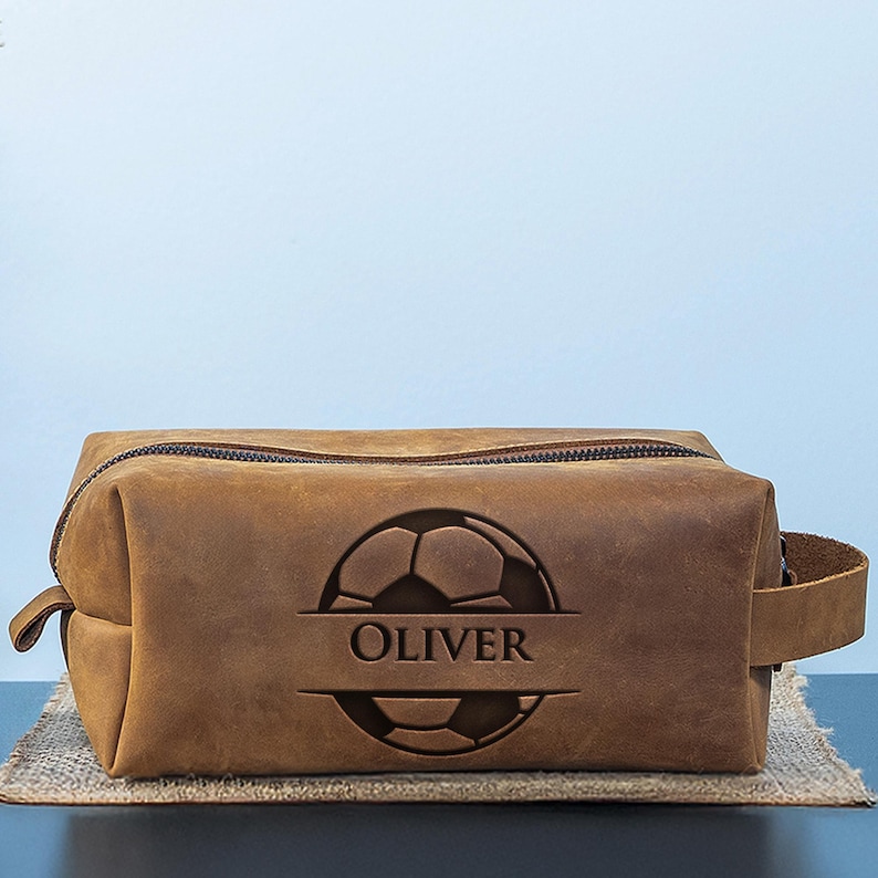 May include: A brown leather wash bag featuring a soccer ball design and the name "OLIVER". The bag has a zip closure and a small handle. The rectangular bag is displayed on a neutral surface. The bag is made of leather.