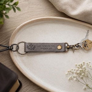 Könnte beinhalten: Ein dunkelgrauer Schlüsselanhänger aus Leder mit einem Metalldruckknopf und einem Karabinerhaken. Das Lederband ist mit einem Kreuzsymbol, Blattzweigen und dem Text "GOD IS WITH YOU JOSHUA 1:9" graviert. Ein silberfarbener Schlüssel ist am Ring befestigt.