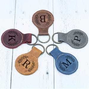 May include: Five leather keychains in various colors: burgundy, brown, tan, gray, and blue. Each keychain features a different initial (B, K, P, R, M) inside a laurel wreath design. The keychains have metal key rings.