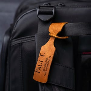 Puede incluir: Una etiqueta de equipaje de cuero marrón con el nombre "PAUL T." y la información de contacto está sujeta a una bolsa de viaje negra. La etiqueta es rectangular con bordes redondeados y se asegura con un lazo. El texto está en una fuente serif.