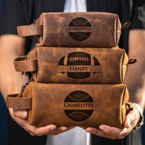 May include: Three brown leather wash bags are stacked. The top and bottom bags feature a basketball design with the name "Charlotte." The middle bag has a football design with the name "Henry." Each bag has a handle.