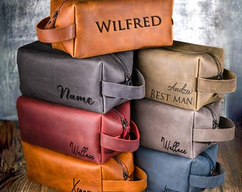 Puede incluir: Una colección de neceseres rectangulares de cuero en varios colores, incluyendo tostado, gris oscuro, rojo y azul. Cada bolsa cuenta con un asa lateral, cierre de cremallera y grabados personalizados como Wilfred, Name, Andzw BEST MAN, Wallace y Xenon.