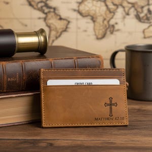 May include: A brown leather cardholder with a cross design and the text "Matthew 42:10". A credit card is visible in one of the slots. The cardholder is on a wooden surface, with a book, a telescope, and a mug in the background.